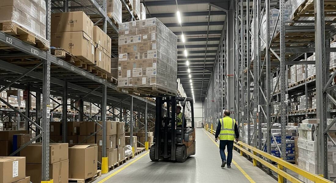Forklift carrying a tall shrink-wrapped load down a warehouse aisle while a pedestrian in a high-visibility vest walks in the same aisle